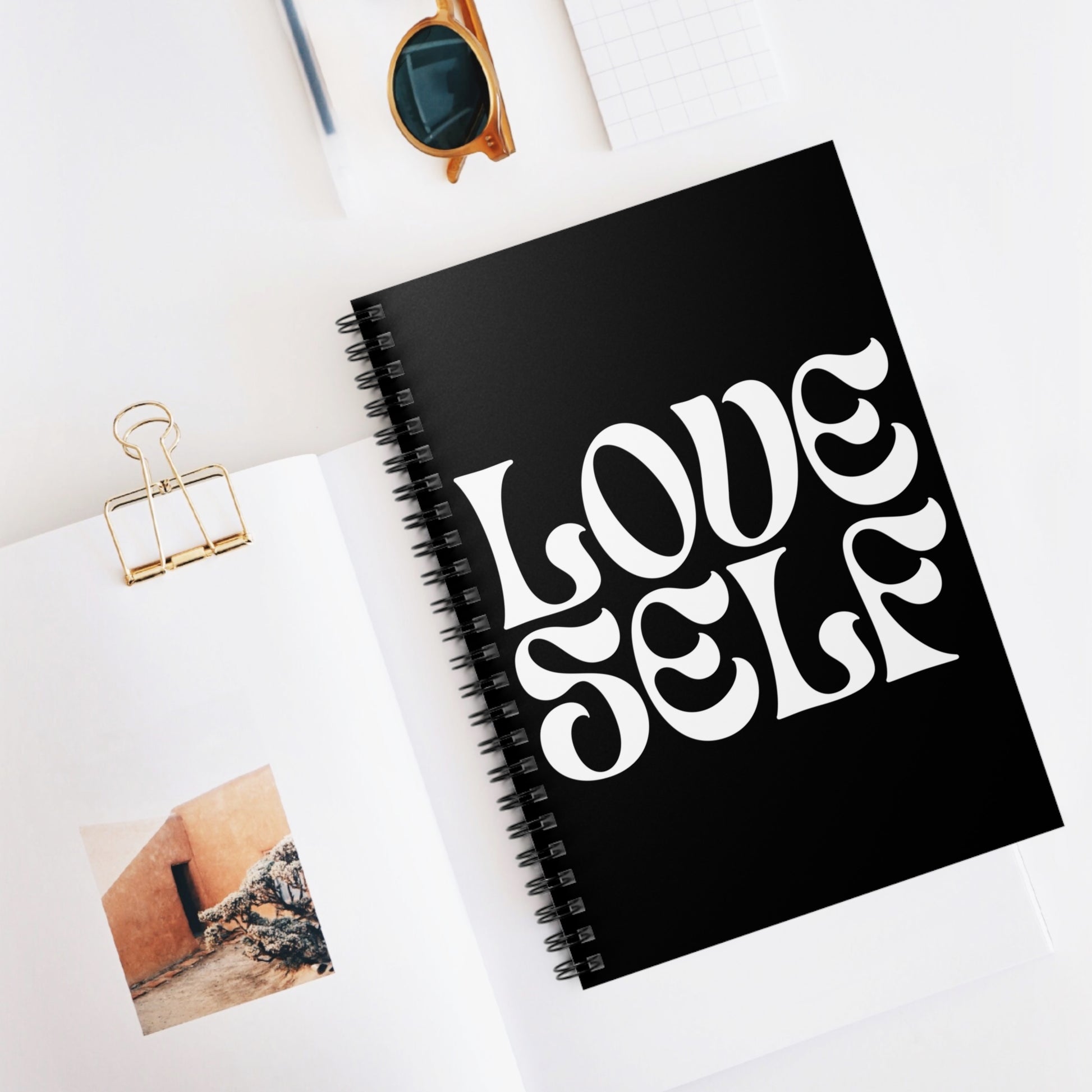 Black notebook with 'LOVE SELF' text on a white surface with sunglasses and a photo clip.