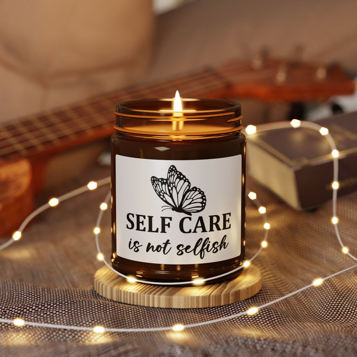 Candle in a jar with a label reading 'Self care is not selfish' on a wooden coaster with fairy lights and a guitar in the background.