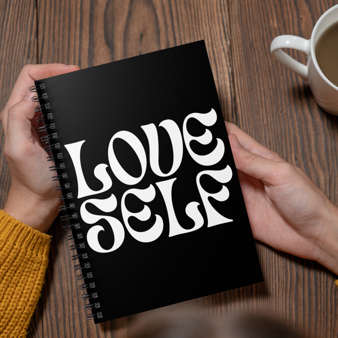 Black notebook with 'LOVE SELF' text held by hands on a wooden table with a cup of coffee.