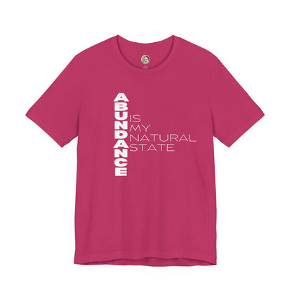 Pink t-shirt with white text 'ABUNDANCE IS MY NATURAL STATE' on a white background