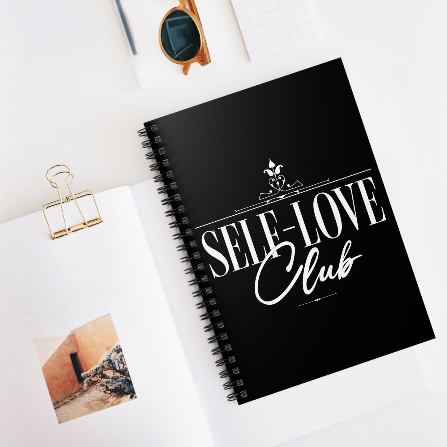 Black notebook with 'Self-Love Club' text on a white surface with sunglasses and a photo clip.